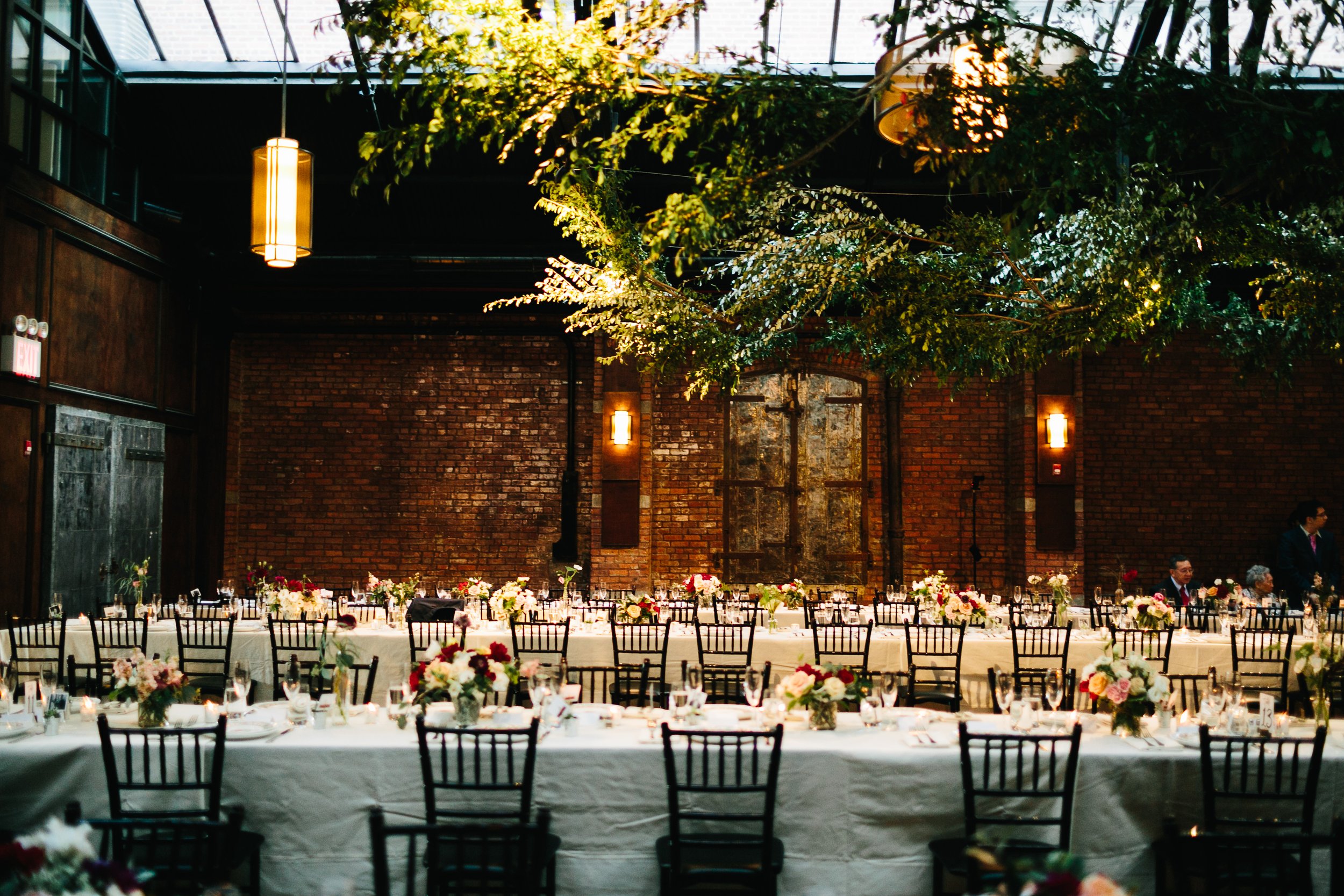 Wedding reception hall with exposed brick, white linens, black chairs, and red flower centerpieces.