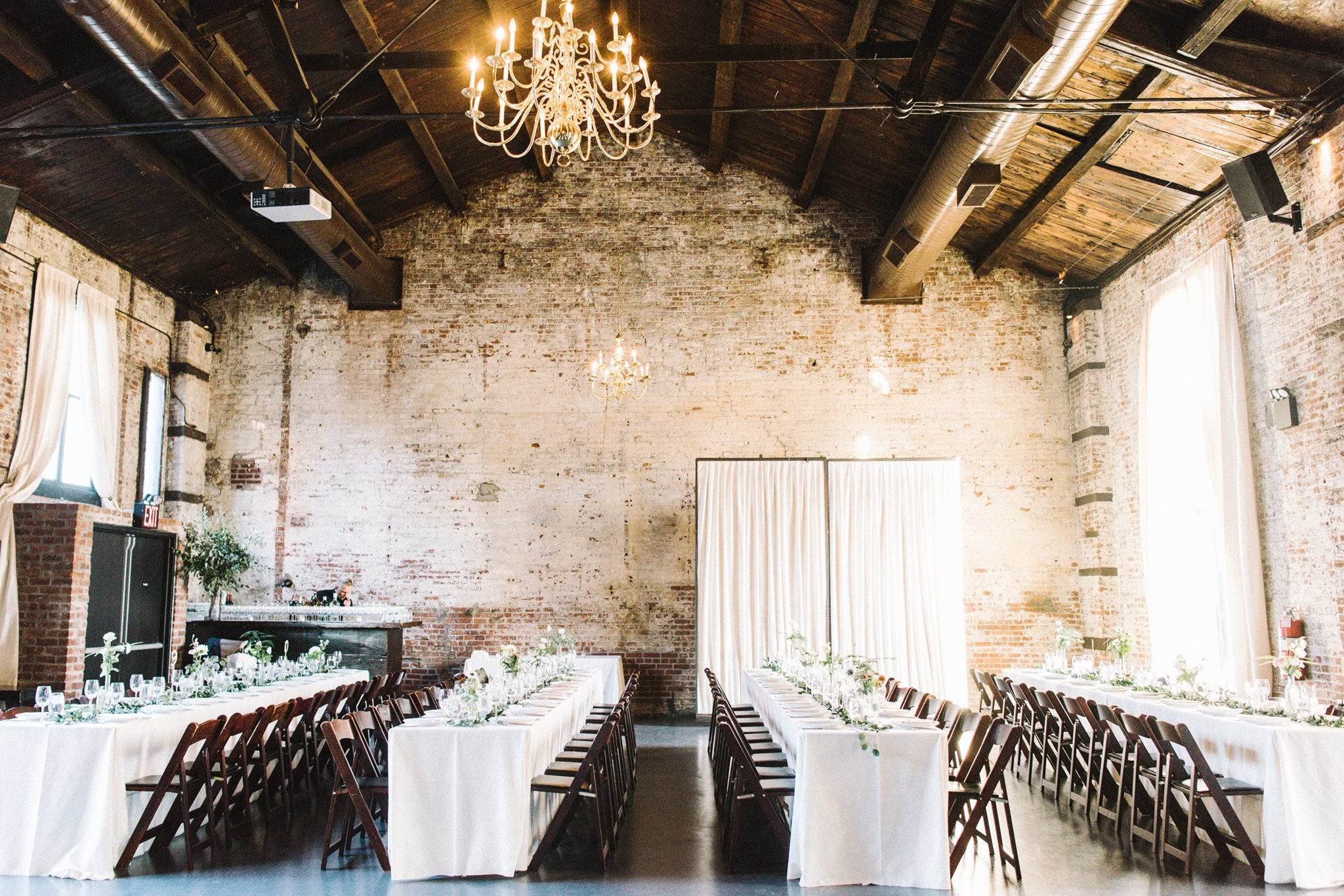 Bright wedding venue with long tables, white flowers, brick walls, and a crystal chandelier.