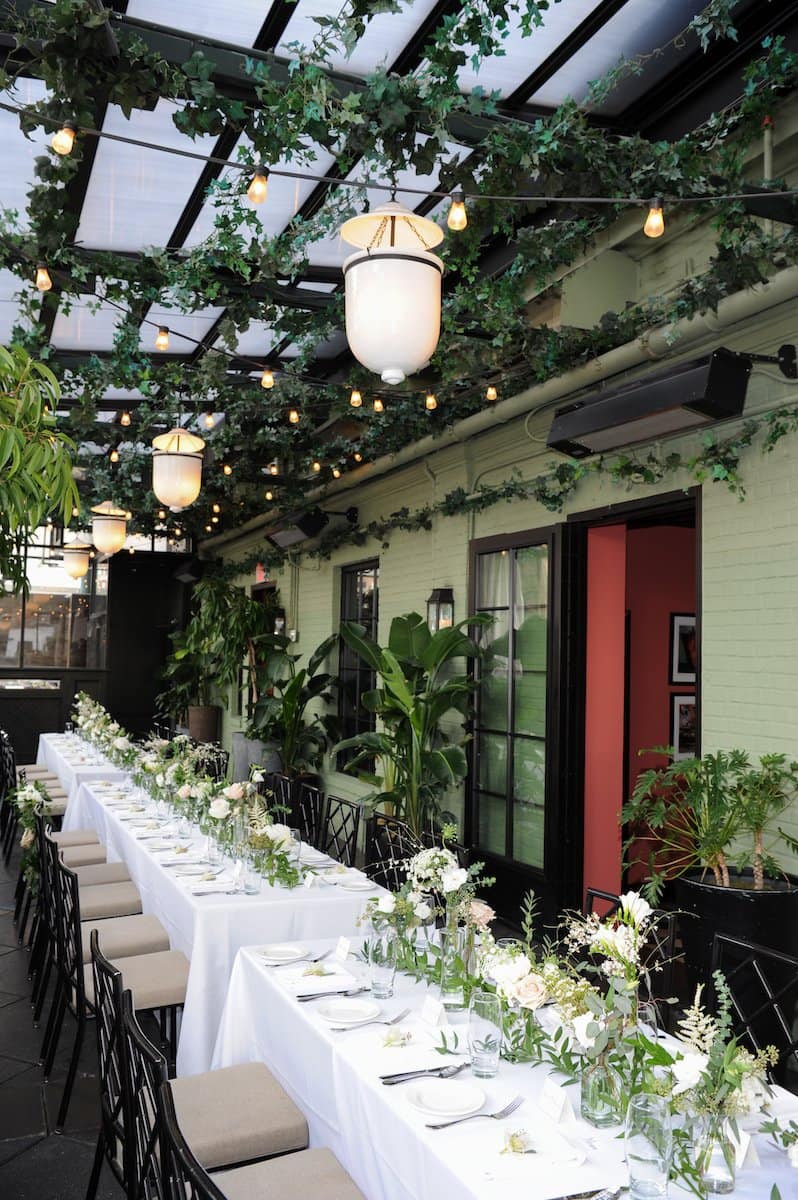 Outdoor dining area with white tables lined up under a ceiling of green foliage and string lights.