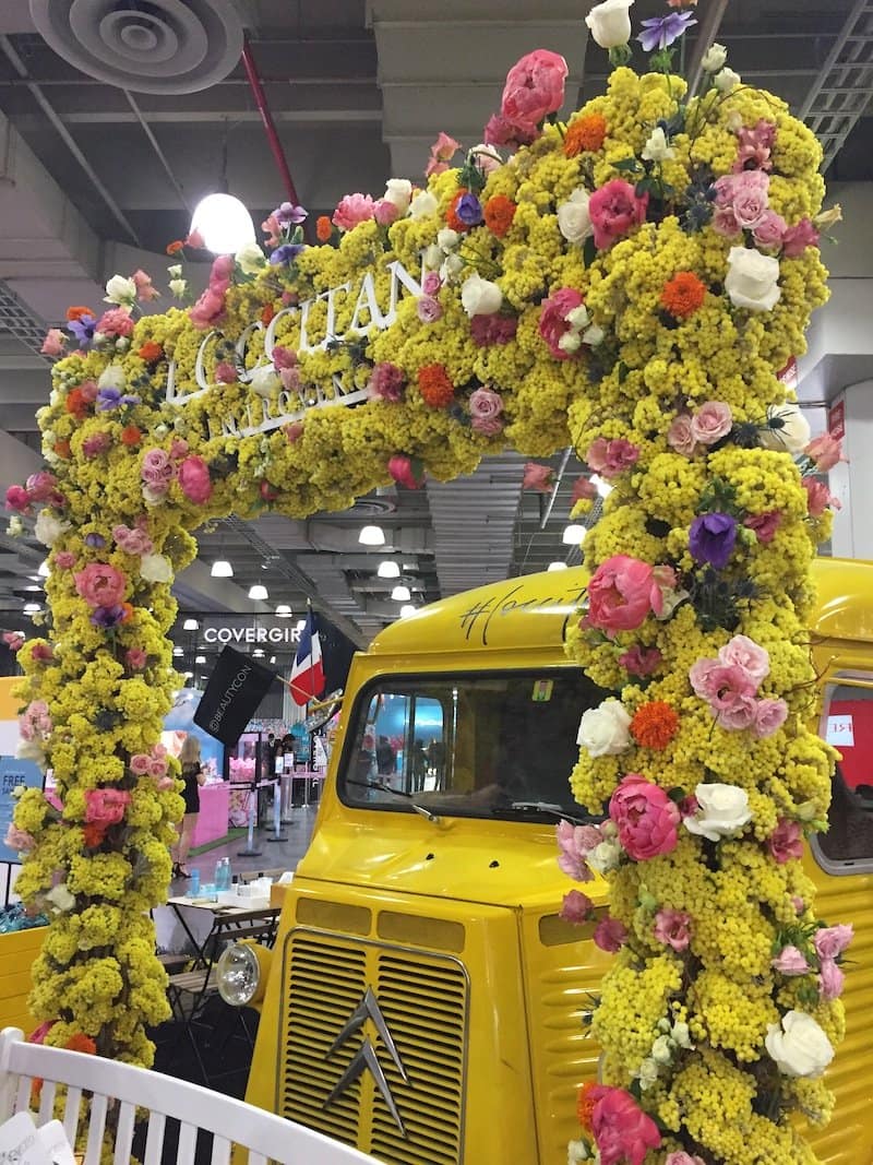 Yellow vintage truck decorated with lush yellow and pink flowers under a floral arch with "L'OCCITANE" signage