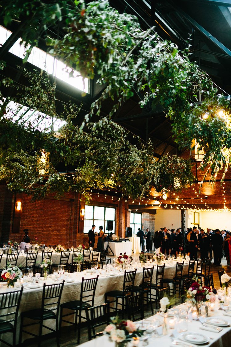 Lively wedding reception with guests, string lights, and greenery overhead in a rustic brick venue.